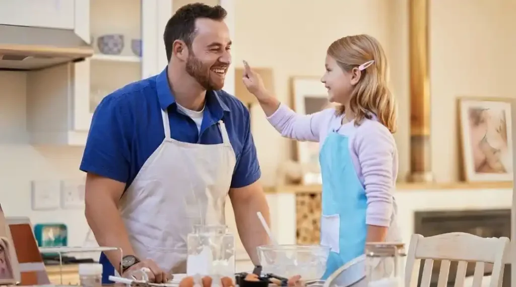 Smiling dad in an apron playfully baking with his daughter, who puts flour on his nose in a warm kitchen setting