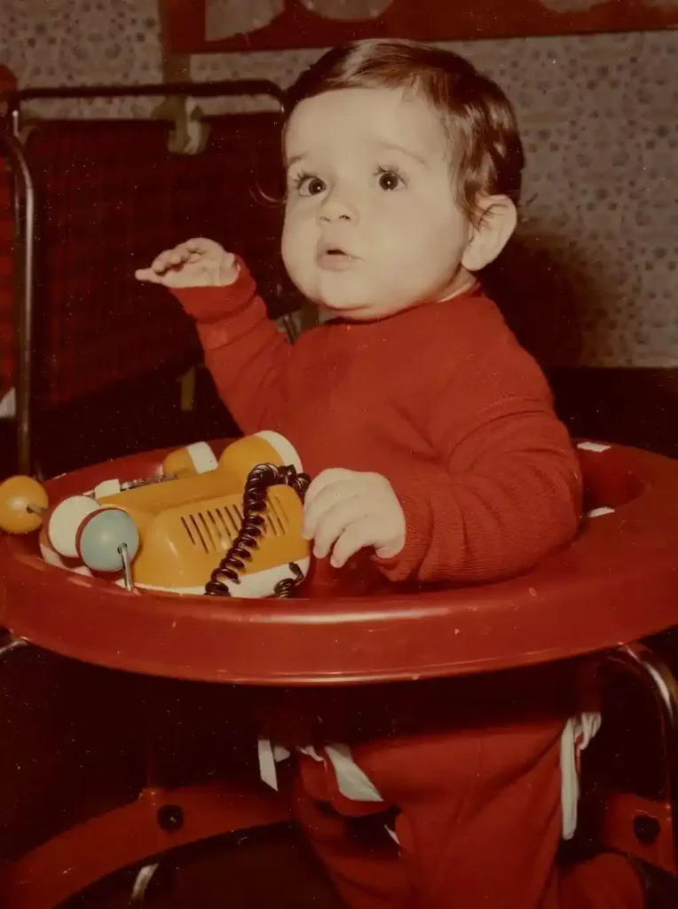 Vintage baby photo of a child in a red outfit sitting in a walker with a toy phone